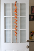 Orange and white crochet wind spinner with green stem and wooden-bead tail hanging against a door.