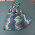 Hand-crocheted Japanese knot bag in teal, purple, green, and cream zigzag colors on a teal background